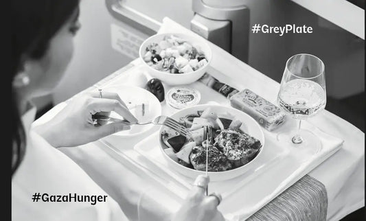 A greyscale photo of a woman dining on a luxury airline meal, with gourmet dishes and a glass of sparkling drink on a white tray table. The hashtags #GreyPlate and #GazaHunger are overlaid, highlighting the contrast between indulgence and crisis.