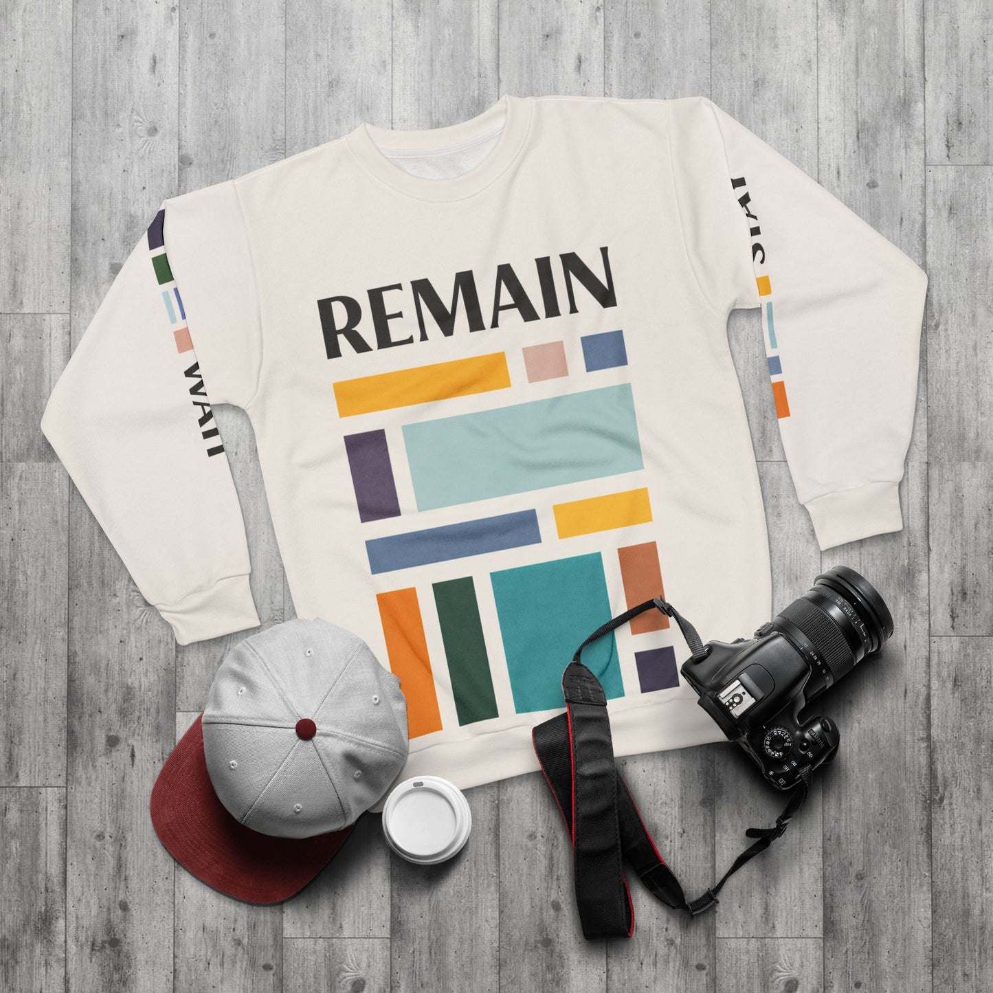 Flat lay of abstract color block sweatshirt with muted teal, mustard, rust and navy geometric print and the word REMAIN, styled with a camera, cap and coffee cup