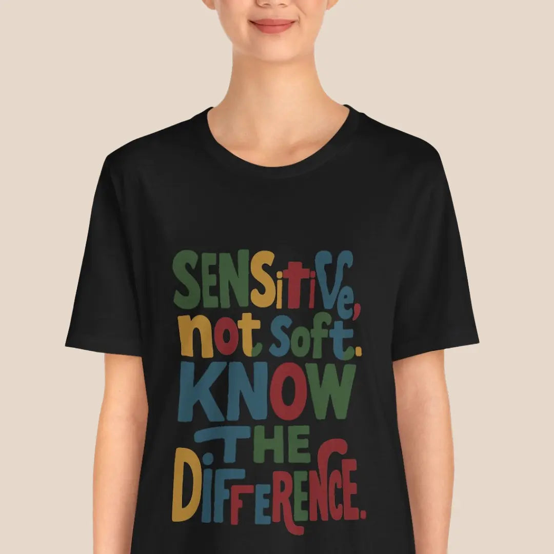 Person wearing a black t-shirt with colorful text "sensitive not soft know the difference" on a beige background