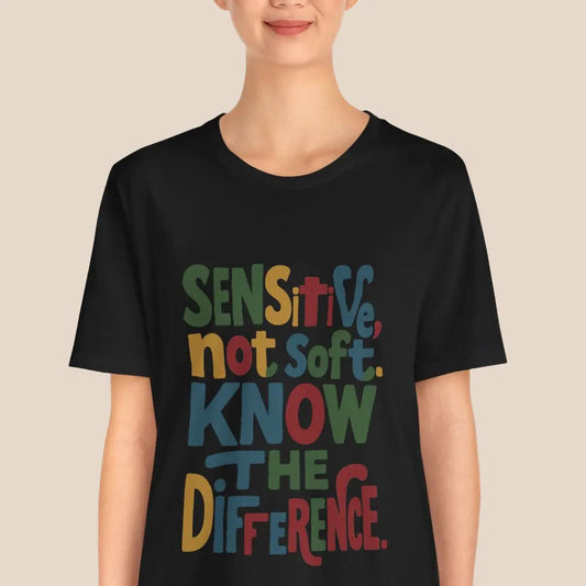 Person wearing a black t-shirt with colorful text "sensitive not soft know the difference" on a beige background