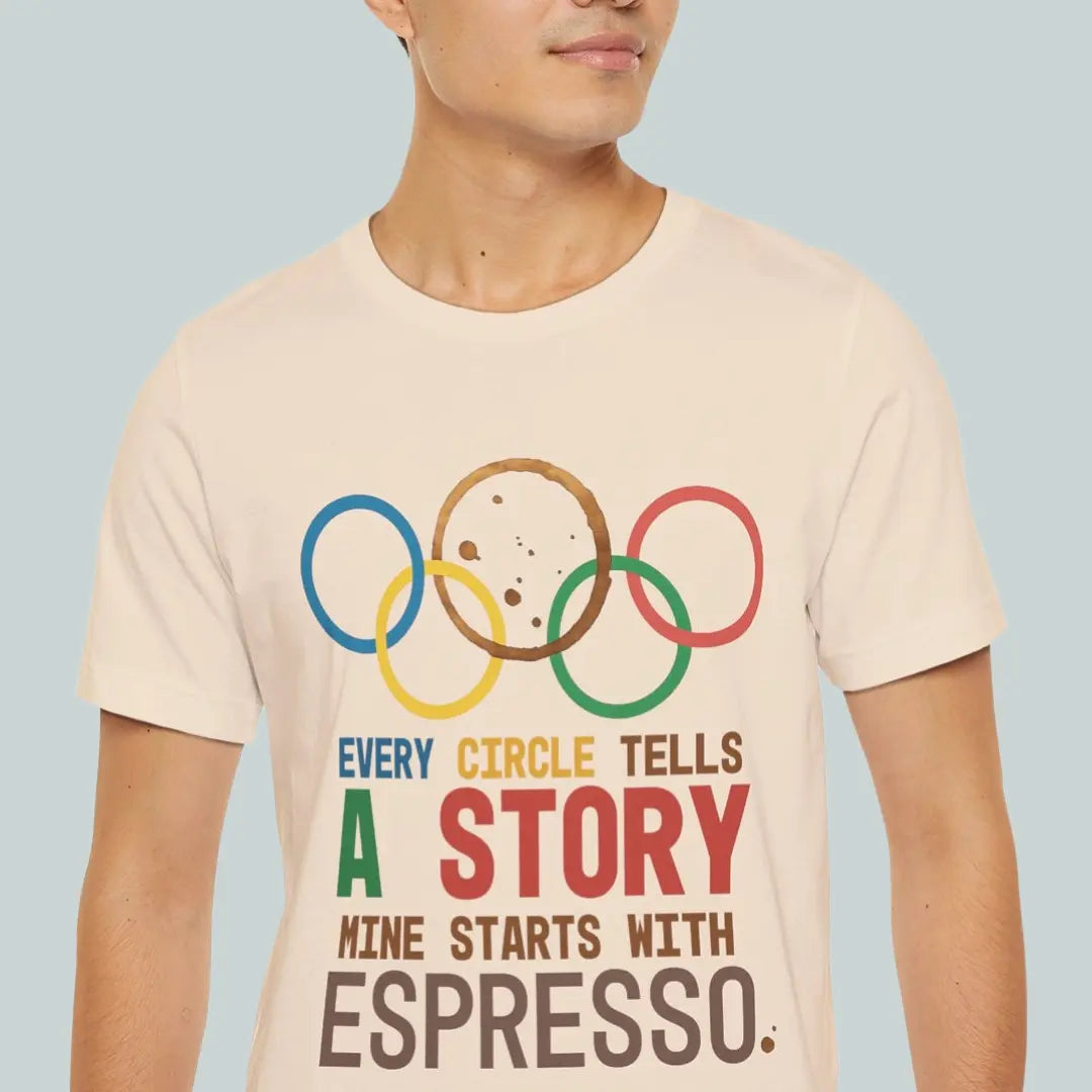 Person wearing a t-shirt with Olympic rings and humorous text every circle tells a story mine starts with espresso, on a plain background