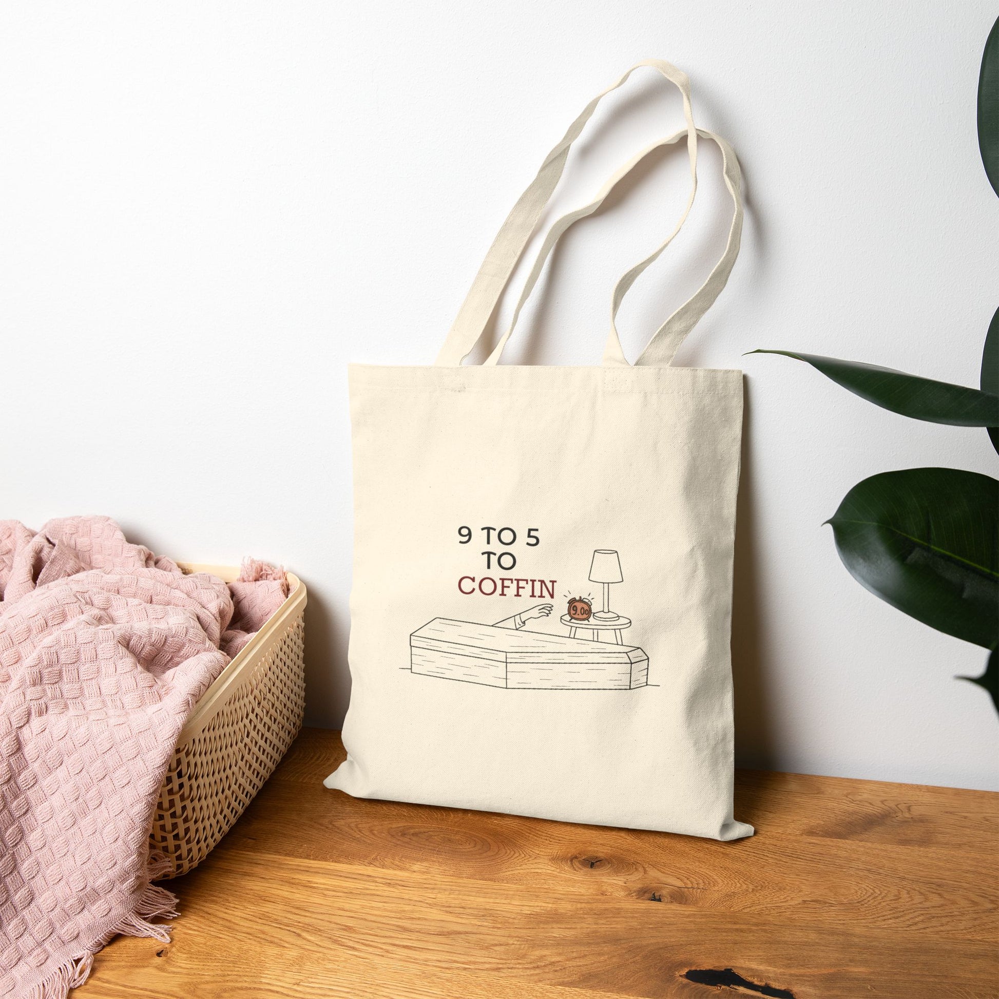 Beige Dead Inside tote bag with 9 TO 5 TO COFFIN typography and coffin illustration for burnout humor for introverts with a plant in the background