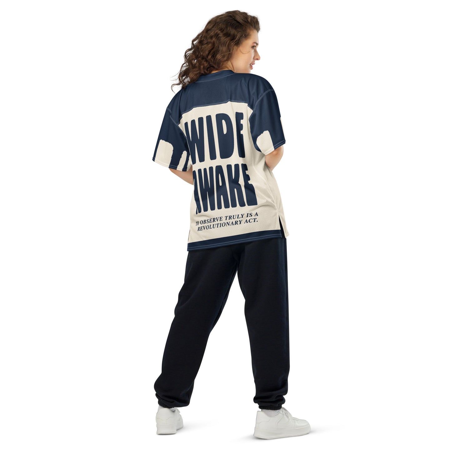 A woman wearing an oversized, all-over-print athletic jersey with bold navy blue typography on the back. The structured block text "WIDE AWAKE" contrasts with the raw, chaotic energy of the front design.