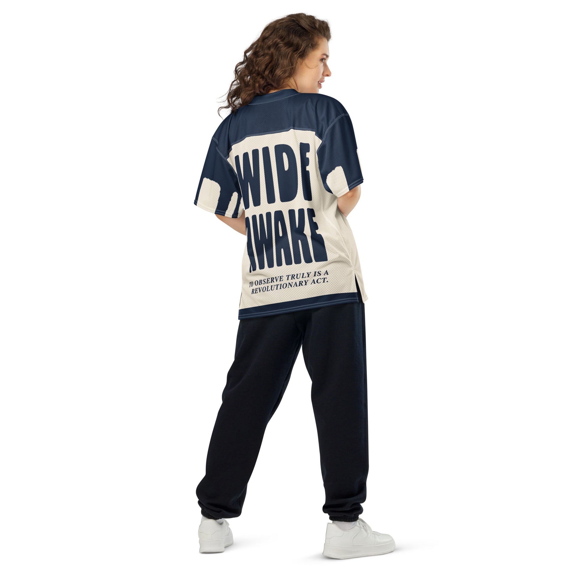 A woman wearing an oversized, all-over-print athletic jersey with bold navy blue typography on the back. The structured block text "WIDE AWAKE" contrasts with the raw, chaotic energy of the front design.