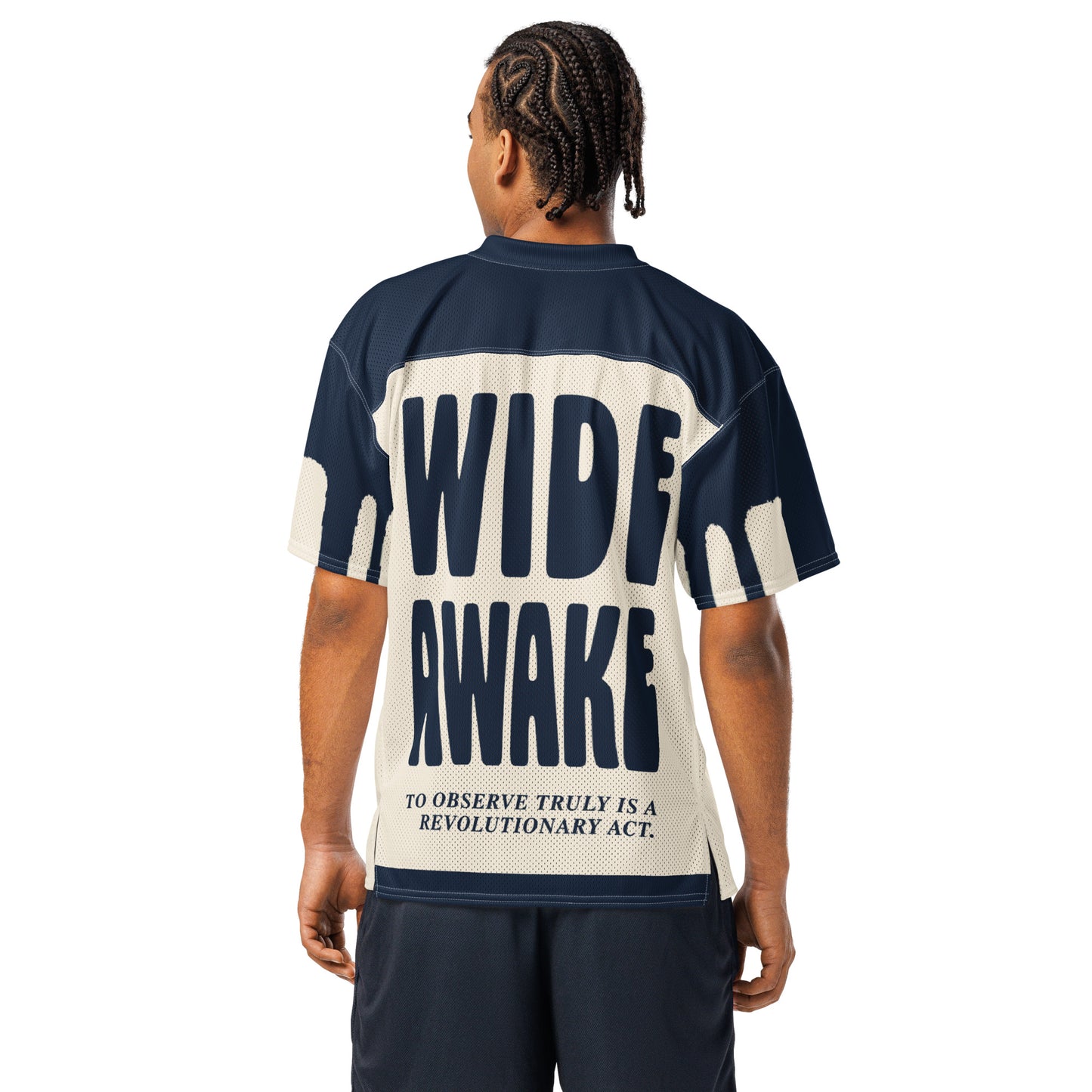 A male model wearing an oversized all-over-print mesh jersey for Text Tease. The back displays bold navy blue "WIDE AWAKE" typography above the text, "To observe truly is a revolutionary act," emphasizing a structured and authoritative aesthetic.