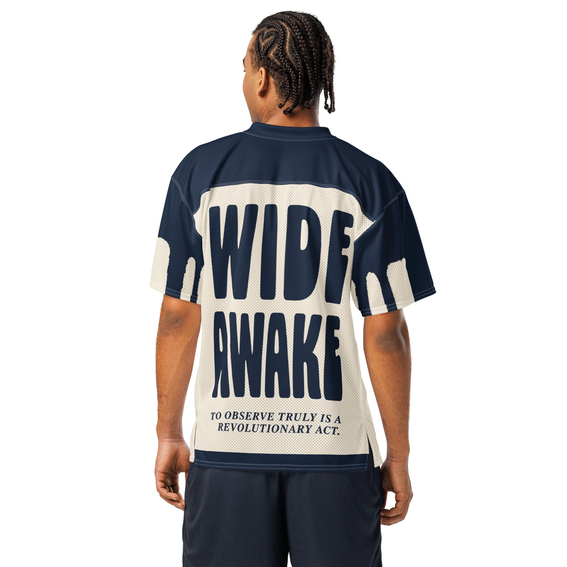 A male model wearing an oversized all-over-print mesh jersey for Text Tease. The back displays bold navy blue "WIDE AWAKE" typography above the text, "To observe truly is a revolutionary act," emphasizing a structured and authoritative aesthetic.