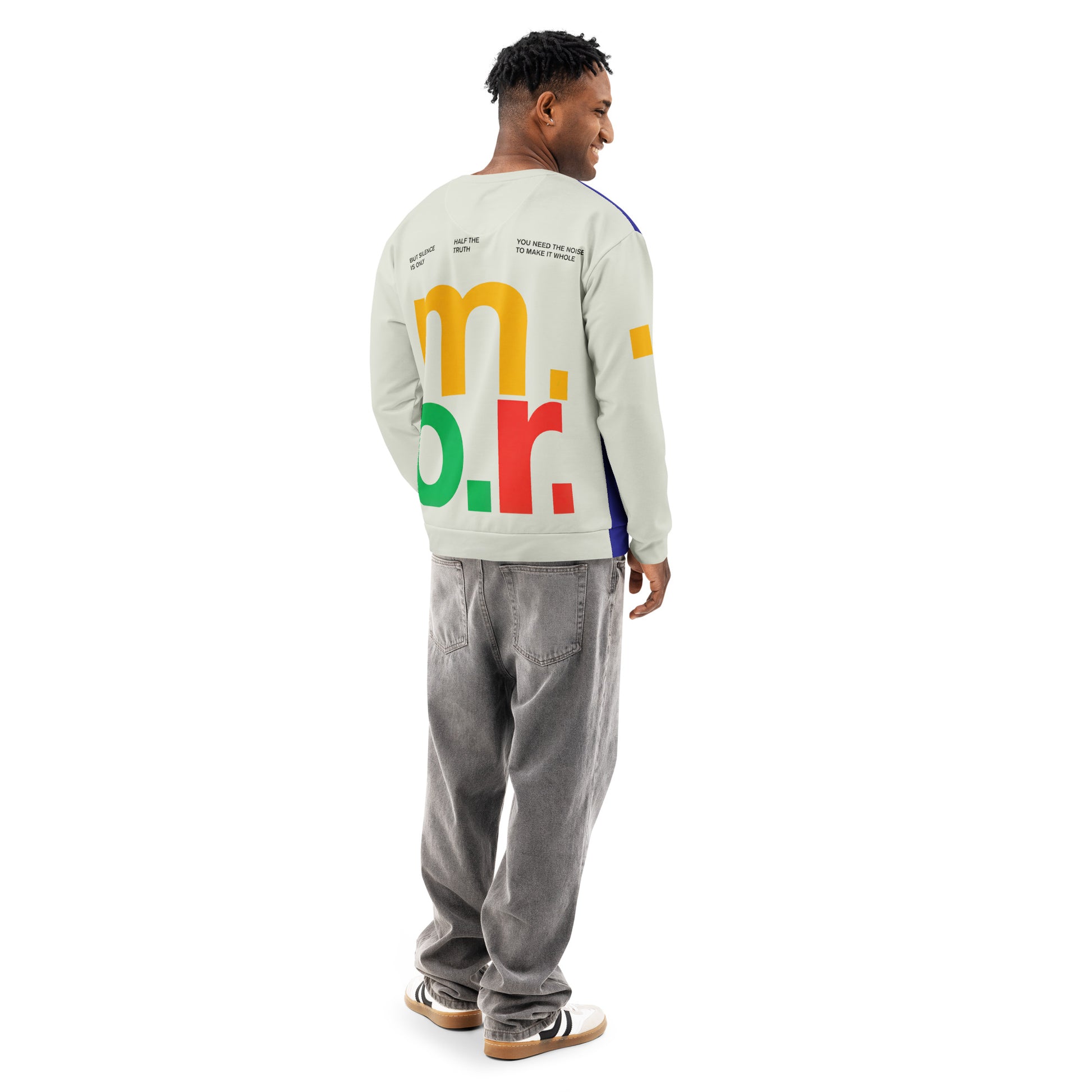 A rear three-quarter view of a male model wearing the Dialectic Crewneck. The design features the vibrant, multi-colored "m.o.r." typography in yellow, green, and red against a light cream-colored fabric, complemented by grey, relaxed-fit denim jeans.