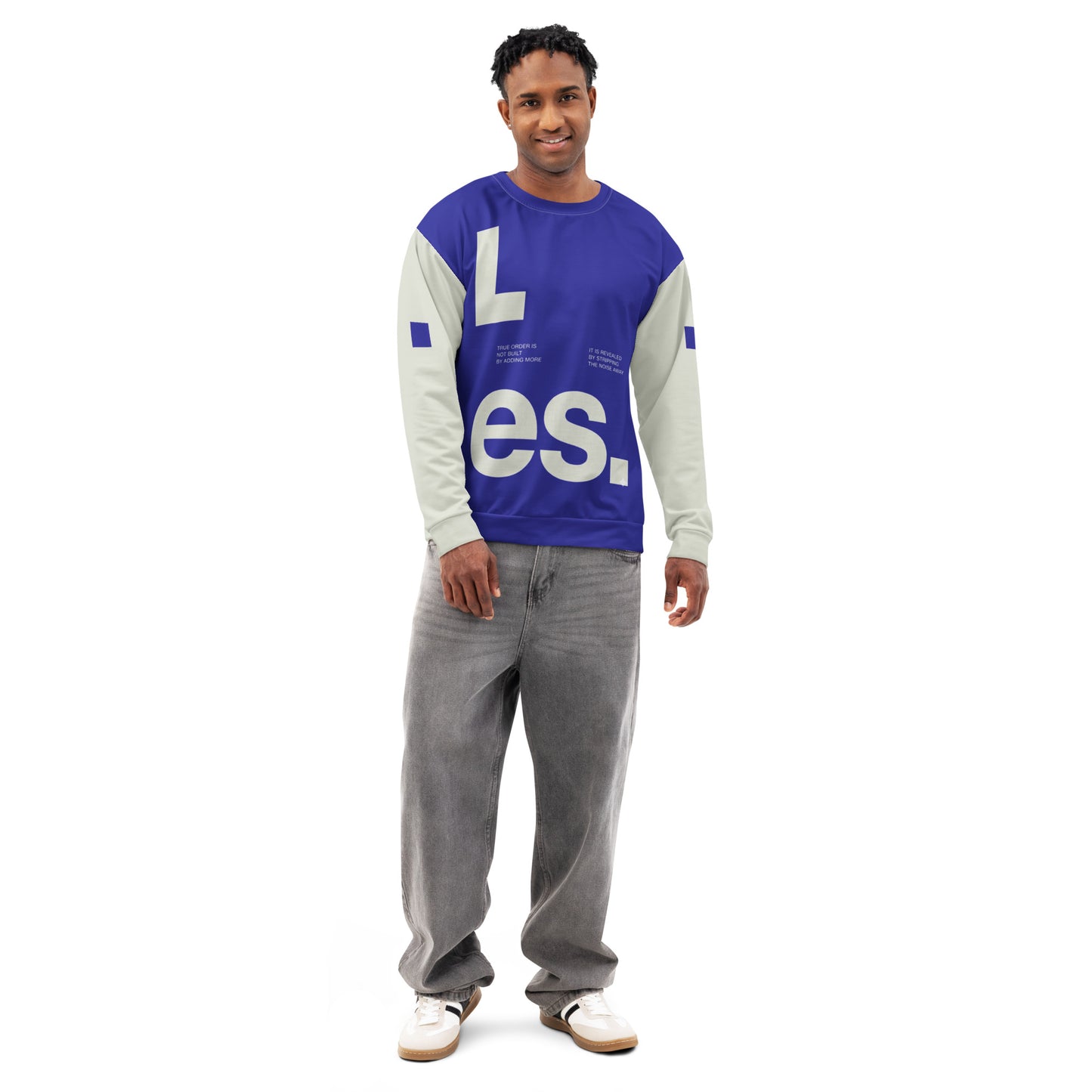 A front-facing, full-length shot of a male model wearing the Dialectic Crewneck. The deep blue, minimalist front panel featuring the bold, white "L.es." graphic is prominently displayed against the light cream-colored sleeves and balanced by a pair of relaxed-fit, grey denim trousers.