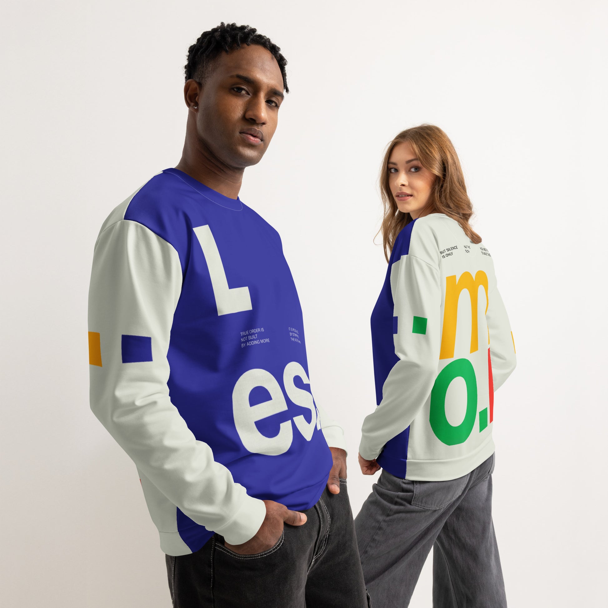 A group lifestyle shot featuring two models wearing the Dialectic Crewneck. The male model in the foreground is seen from a three-quarter side view, highlighting the deep blue front panel with ivory "L.es." typography. The female model in the background is seen from a three-quarter rear view, showcasing the vibrant, multi-colored "m.o.r." graphic in yellow, green, and red against the cream-colored fabric. Both models are styled with dark grey, relaxed-fit denim.